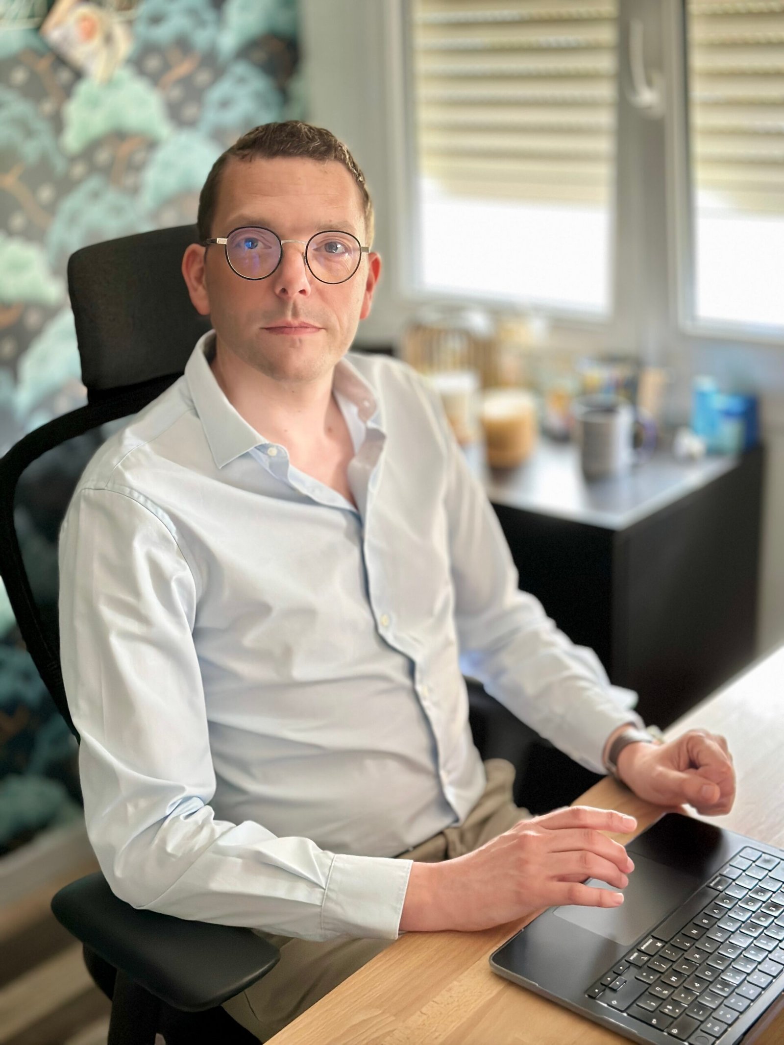 Un homme portant des lunettes et une chemise blanche est assis à un bureau devant un ordinateur portable. L'arrière-plan montre une fenêtre avec des stores et des étagères.