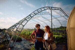 Un agriculteur et une femme en tenue professionnelle se tiennent à l'intérieur d'une serre et regardent ensemble un bloc-notes.