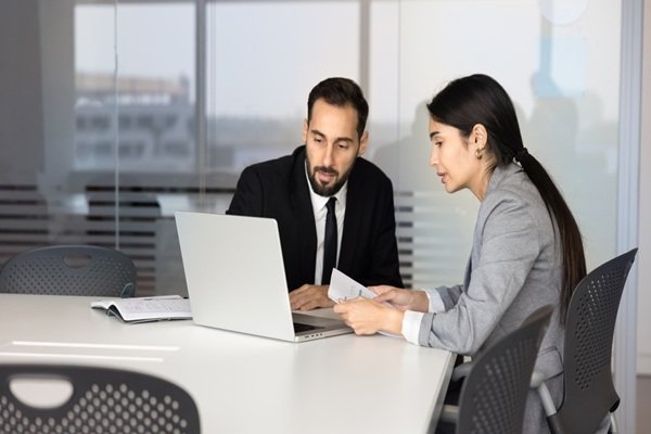 Un homme et une femme en tenue professionnelle, assis à une table, discutent de leur travail sur un ordinateur portable.