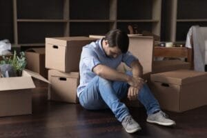 Un homme assis par terre, le regard baissé, entouré de cartons de déménagement.
