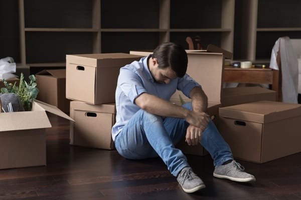 Un homme assis par terre, le regard baissé, entouré de cartons de déménagement.