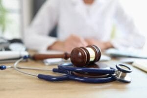 Le marteau d'un juge posé sur un stéthoscope, sur un bureau en bois, avec en arrière-plan une personne vêtue d'une blouse blanche.