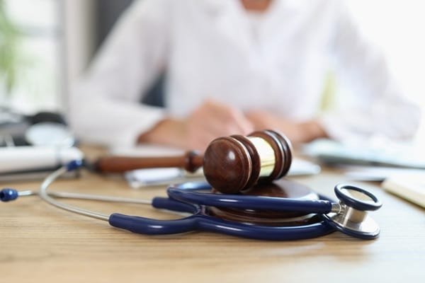 Le marteau d'un juge posé sur un stéthoscope, sur un bureau en bois, avec en arrière-plan une personne vêtue d'une blouse blanche.