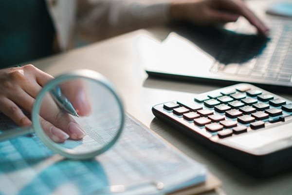Une personne utilisant une loupe pour examiner un document, avec une calculatrice et un ordinateur portable à proximité.