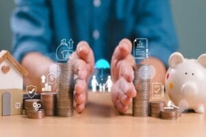 Des mains formant une coupe renferment des piles de pièces, une petite maquette de maison et une tirelire, ainsi que des icônes numériques symbolisant l'assurance et la protection financière.