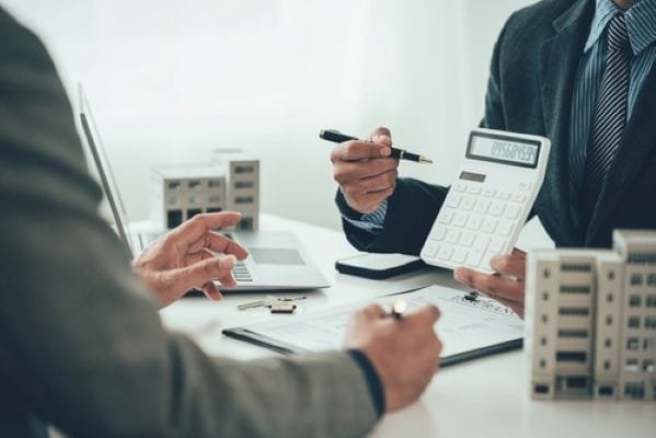Deux personnes en tenue professionnelle examinant des documents et une calculatrice, assises à un bureau sur lequel sont posées des maquettes de bâtiments.