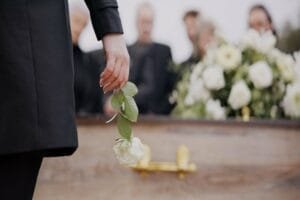 Une personne vêtue d'un manteau noir, tenant une rose blanche au-dessus d'un cercueil en bois lors d'un enterrement.