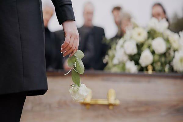 Une personne vêtue d'un manteau noir, tenant une rose blanche au-dessus d'un cercueil en bois lors d'un enterrement.