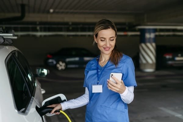 Une femme vêtue d'une blouse bleue recharge un véhicule électrique dans un parking couvert tout en consultant son smartphone.