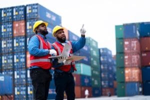 Deux dockers, coiffés de casques de chantier et vêtus de gilets de sécurité, se tiennent dans une cour où s'empilent des conteneurs ; l'un d'eux pointe du doigt vers le haut tout en regardant un ordinateur portable.