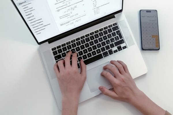 Une personne qui tape sur un ordinateur portable, tandis qu'une application de calculatrice est ouverte sur un smartphone posé à proximité.