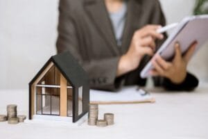 Une petite maquette de maison trône sur une table, à côté de piles de pièces de monnaie, tandis qu'à l'arrière-plan, une personne utilise une tablette.