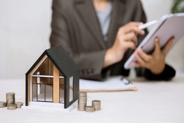 Une petite maquette de maison trône sur une table, à côté de piles de pièces de monnaie, tandis qu'à l'arrière-plan, une personne utilise une tablette.